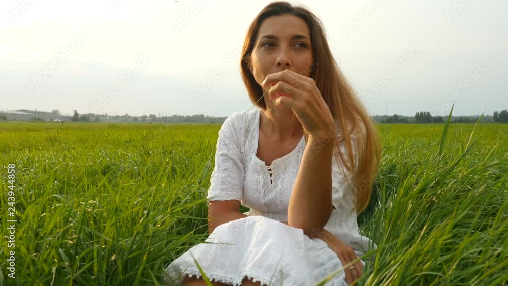 Portrait happy young woman on a summer walk in field. Cute girl sitting on the green grass and dreaming. Beautiful young woman enjoying nature in summer evening. Grassy summerfield Portrait happy young woman on a summer walk in field. Cute girl sitting on the green grass and dreaming. Beautiful young woman enjoying nature in summer evening. Grassy summerfield