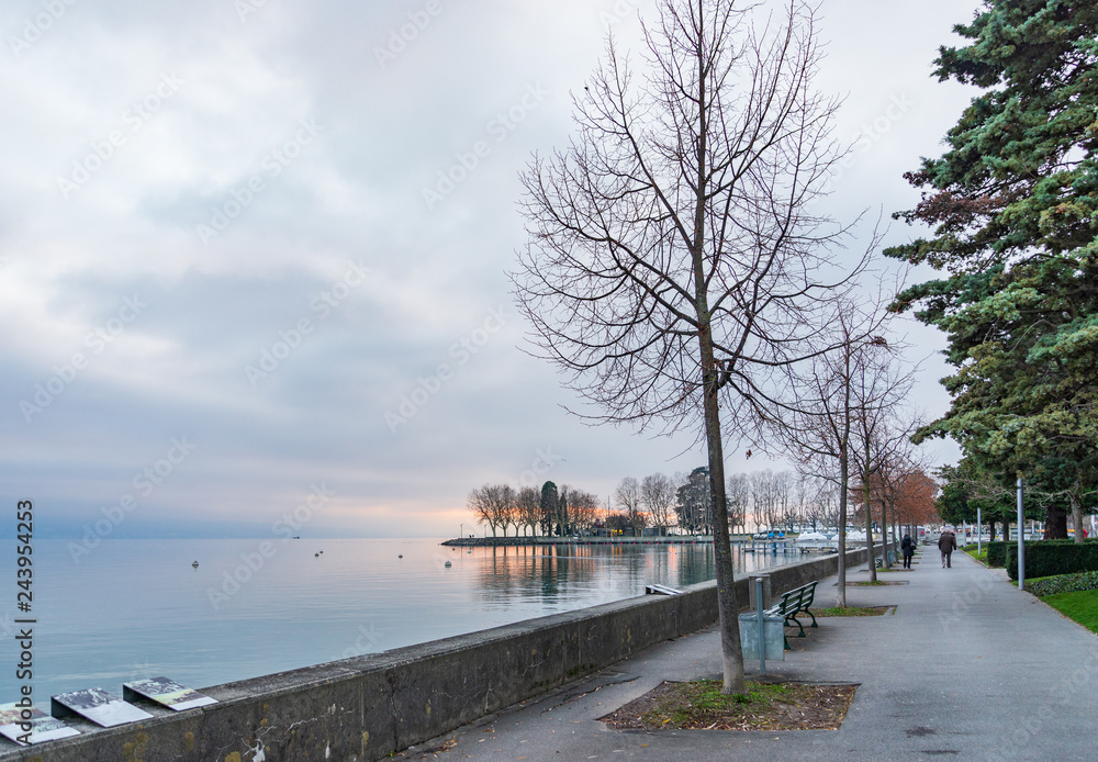 Outdoor scenery of beautiful tranquil natural walkway and promenade along lakeside of lake Geneva and background of misty, cloudy and twilight sky over water in Lausanne, Switzerland in winter.