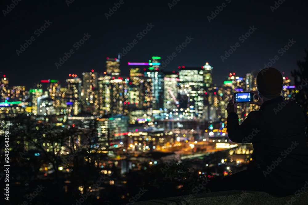 Man Sitting on Ledge Taking Phone Pics of Blurry City Lights Background for Social Media