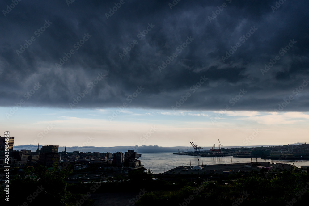 A dark storm cloud over a Saint John city in New Brunswick, Canada. White cloud and blue sky visible in the distance. City is very dark because of the cloud.