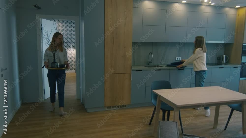 Two young female designers decorating table in modern dining room in luxury apartment: woman bringing vase with twigs while her colleague putting blue table runner Two young female designers decorating table in modern dining room in luxury apartment: woman bringing vase with twigs while her colleague putting blue table runner