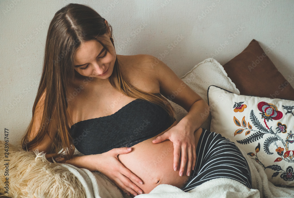 Naklejka premium Attractive pregnant woman is sitting in bed and holding her belly. Last months of pregnancy.
