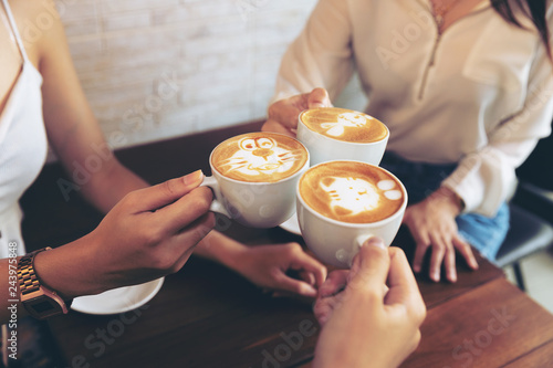 Group of friends cheers with latae cup in cafe bar with phone on table sitting intdoor at cafe - Young girl group having fun drinking together.