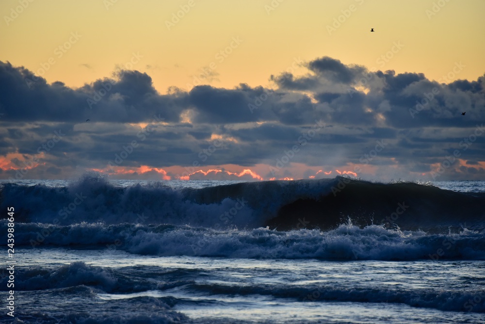 Storm on the sea at sunset. Blue sky with dark clouds. The light of the sun on the horizon. Big waves roll ashore. Splashes and mist over the wave.
