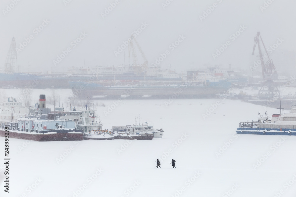 Big river crafts on the winter parking. The ships are frozen in ice. On a background port cranes. Two men of fishermen go through a blizzard on the surface of ice