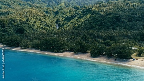 aerial drone image of a south pacific village on a remote island with sandy beach shore and lush tropical rainforest jungle with waterfall in the background © simanlaci