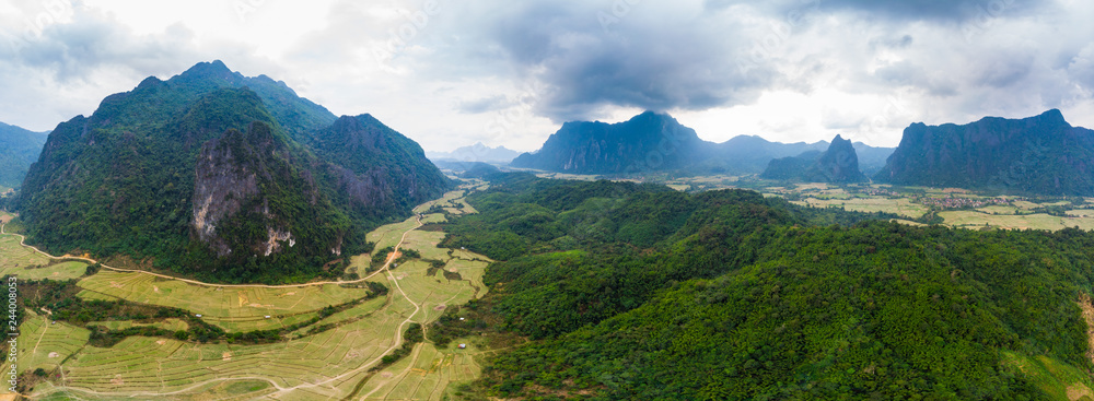 Aerial: Vang Vieng backpacker travel destination in Laos, Asia. Dramatic sky over scenic cliffs and rock pinnacles, rice paddies valley, stunning landscape.