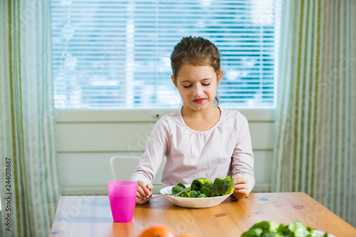 Cute girl eating spinach and broccoli at the table. Child doesn't want to eat, refuses eating, making faces. Healthy food concept. 