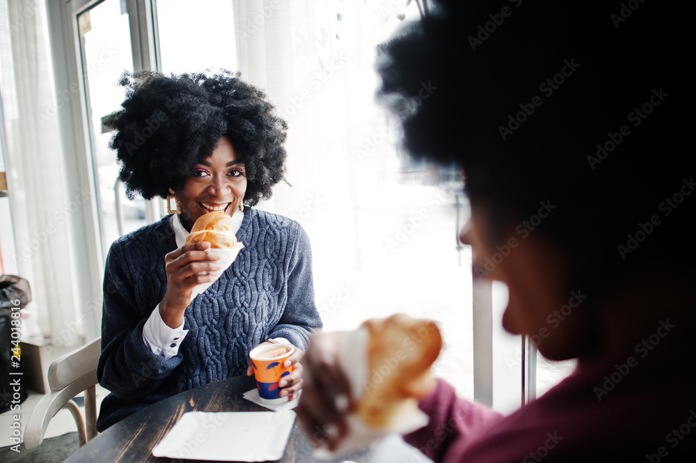 Two curly hair african american woman wear on sweaters sits at table cafe , eat croissant and drink tea.