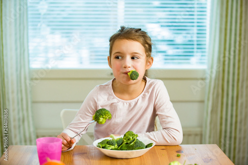Cute girl eating spinach and broccoli at the table. Smiling and laughing happily. Healthy food concept. 