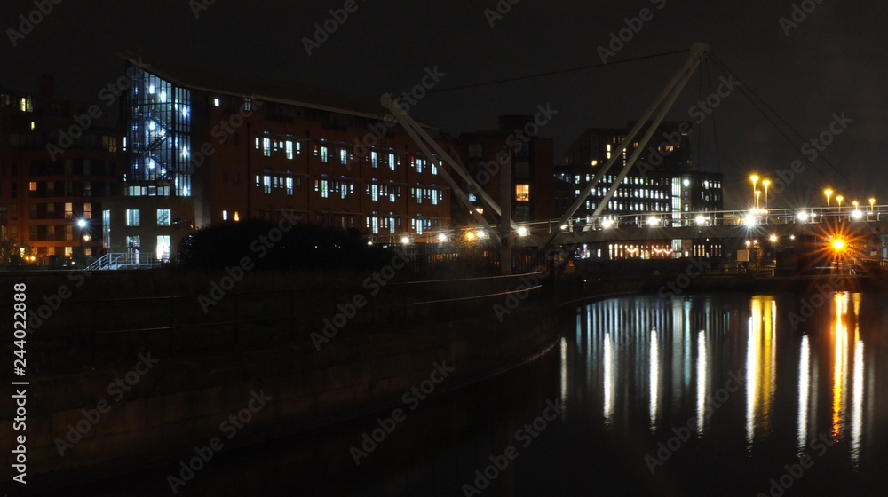 a cityscape view of the canal entrance to the clarence dock area of leeds with a pedestrian bridge crossing the water with reflections of lights and buildings against a night sky with clouds