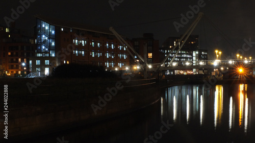 a cityscape view of the canal entrance to the clarence dock area of leeds with a pedestrian bridge crossing the water with reflections of lights and buildings against a night sky with clouds © Philip J Openshaw 