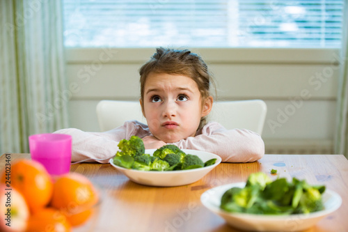 Cute girl eating spinach and broccoli at the table. Child doesn't want to eat, refuses eating, making faces. Healthy food concept. 