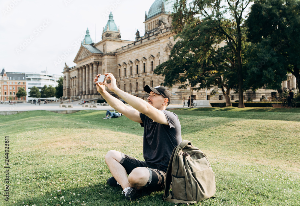 Φωτογραφία A young male tourist or student sits on the grass outside in Leipzig, Germany, talking on video or using a mobile phone to chat or take pictures