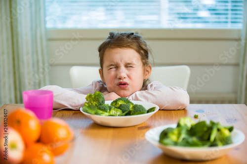 Cute girl eating spinach and broccoli at the table. Child doesn't want to eat, refuses eating, making faces. Healthy food concept. 