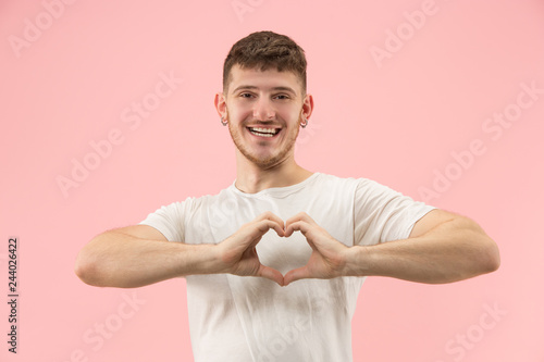 Attractive guy is making a heart shape symbol with his fingers. He is smiling because he loves. Isolated on a pink studio background