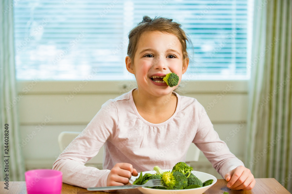 Cute girl eating spinach and broccoli at the table. Smiling and laughing happily. Healthy food concept. Cute girl eating spinach and broccoli at the table. Smiling and laughing happily. Healthy food concept.