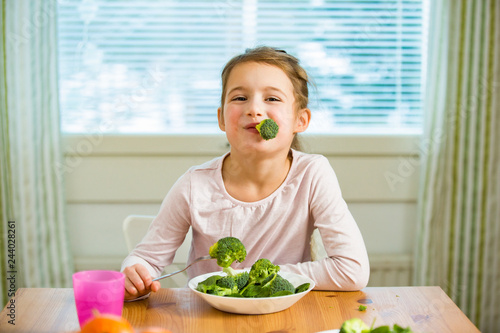 Cute girl eating spinach and broccoli at the table. Smiling and laughing happily. Healthy food concept. 