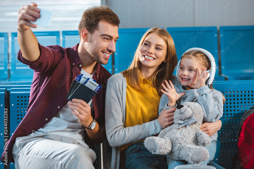 cheerful dad holding passports with air tickets and taking selfie with smiling wife and daughter in airport