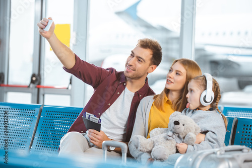 Fotografie cheerful dad taking selfie and smiling near wife and daughter showing tongue in