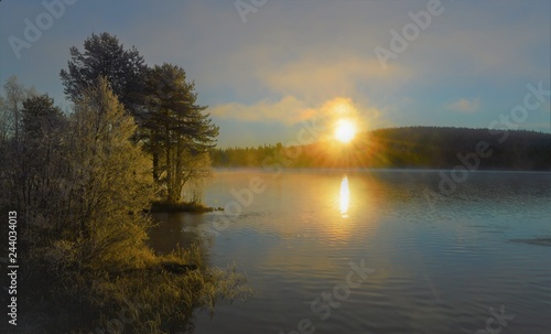 Autumn time in Northern Finland. Cold, bright, beautiful morning. Frost-covered trees, rising sun, misty lake clouds, reflection.