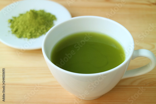 Closed Up a Cup of Hot Matcha Green Tea Served on Wooden Table with Blurry Plate of Matcha Tea Powder in Background