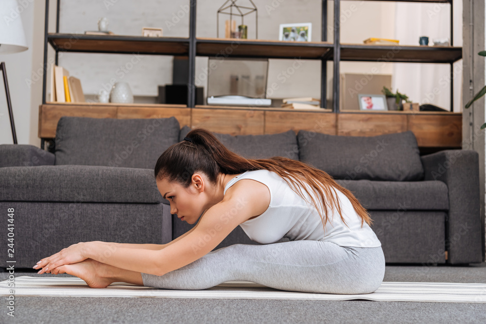 Woman practicing seated forward fold pose on fitness mat at home in living room Woman practicing seated forward fold pose on fitness mat at home in living room