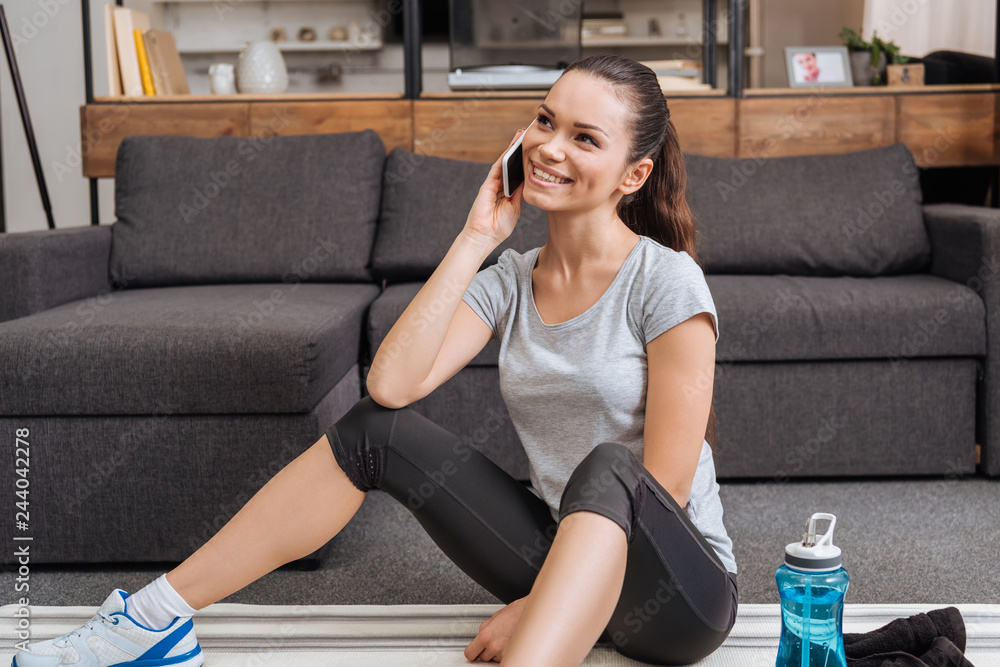 attractive smiling sportswoman sitting on fitness mat and talking on smartphone at home attractive smiling sportswoman sitting on fitness mat and talking on smartphone at home