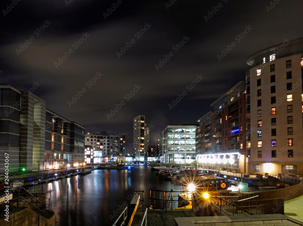 Clarence dock leeds at night with moored barges and moonlit clouds over brightly illuminated waterside buildings reflected in the harbour