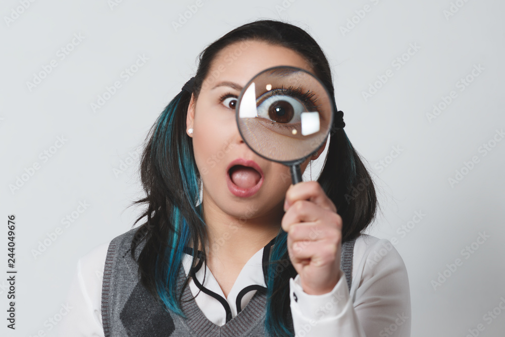 Portrait of a funny young woman looking at the camera through magnifying glass on gray background Portrait of a funny young woman looking at the camera through magnifying glass on gray background