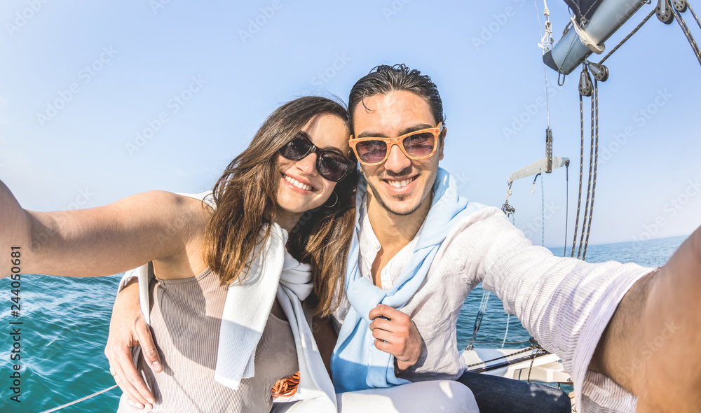 Young lover couple taking selfie on sailing boat tour around the world - Love concept at jubilee party cruise on luxury sailboat experience with happy boyfriend and girlfriend - Warm bright sun filter