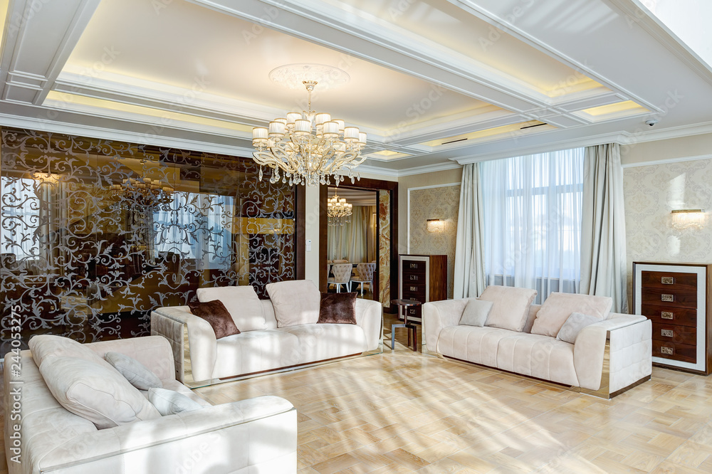 Fototapeta premium Classic interior of living room in brown, white, beige, with glasses wall and crystal chandelier in the center of ceiling. Three big and cozy sofas with pillows for big family.