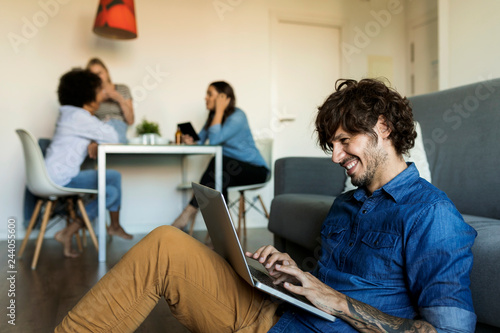 Smiling man sitting on floor using laptop with friends in background