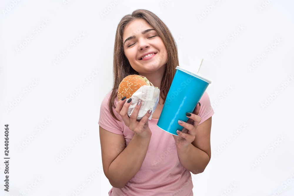 Cheerful and happy young woman hold burger and cup of coca-cola in hands. She looks amazing. Isolated on white background. Cheerful and happy young woman hold burger and cup of coca-cola in hands. She looks amazing. Isolated on white background.