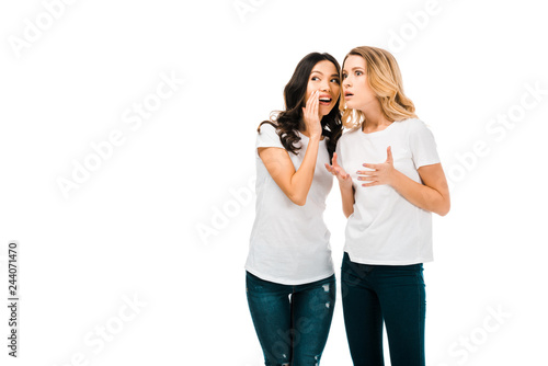 smiling young woman whispering something to shocked girlfriend isolated on white