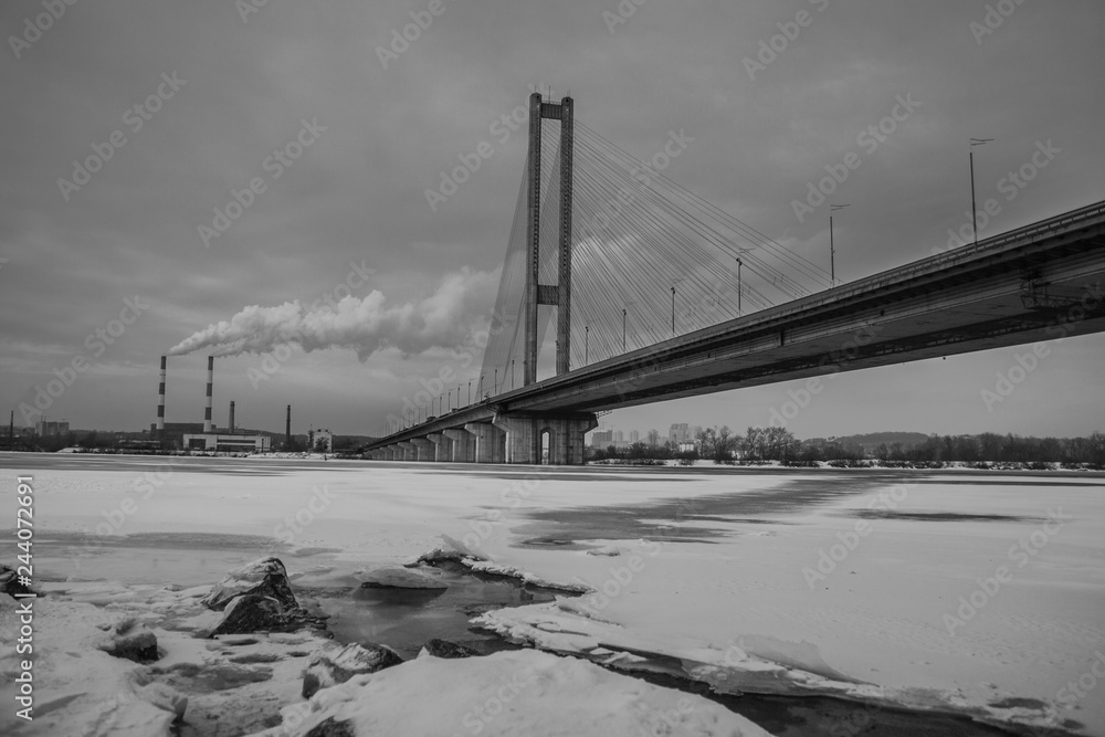 Bridge over the winter frozen river on the background of industrial smoking pipes. Cityscape