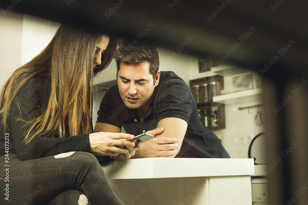 A young couple looking at the cell phone in the kitchen. Concept of technology