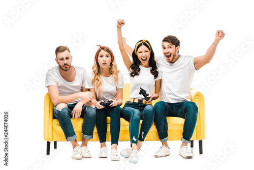 emotional young friends sitting on sofa and playing with joysticks isolated on white