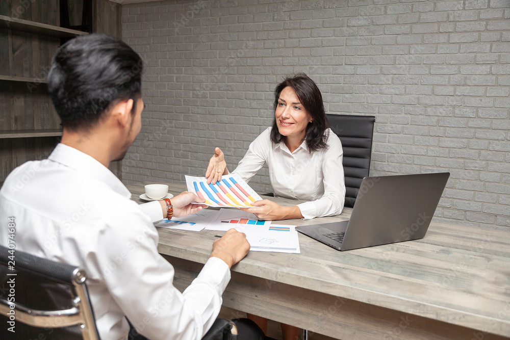 Attractive aged business woman, teacher or mentor coach speaking to young Asian businessman provides recruitment and employment services show graph of progress. Startup and business planning concept. Attractive aged business woman, teacher or mentor coach speaking to young Asian businessman provides recruitment and employment services show graph of progress. Startup and business planning concept.
