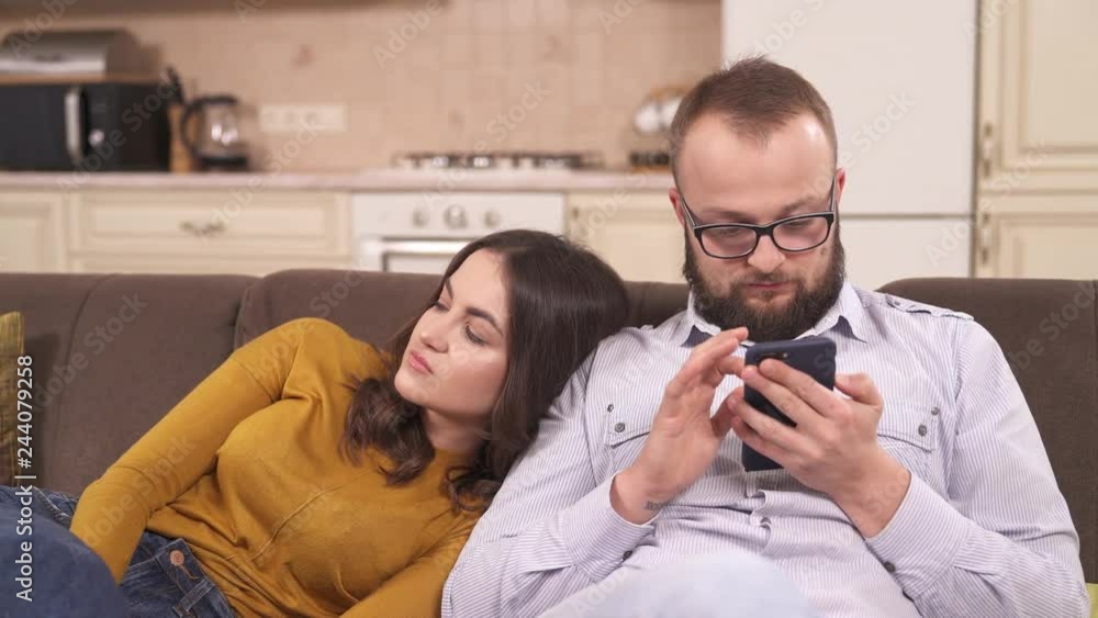 A pretty brunette leans her head against a bearded man in glasses sitting on the sofa and scrolling the phone. She sighs and seems to be bored A pretty brunette leans her head against a bearded man in glasses sitting on the sofa and scrolling the phone. She sighs and seems to be bored