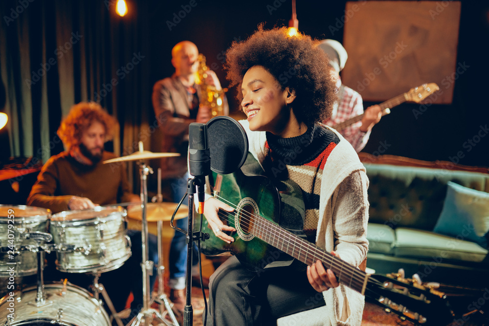 Fototapeta premium Mixed race woman singing and playing guitar while sitting on chair with legs crossed. In background drummer, saxophonist and bass guitarist.