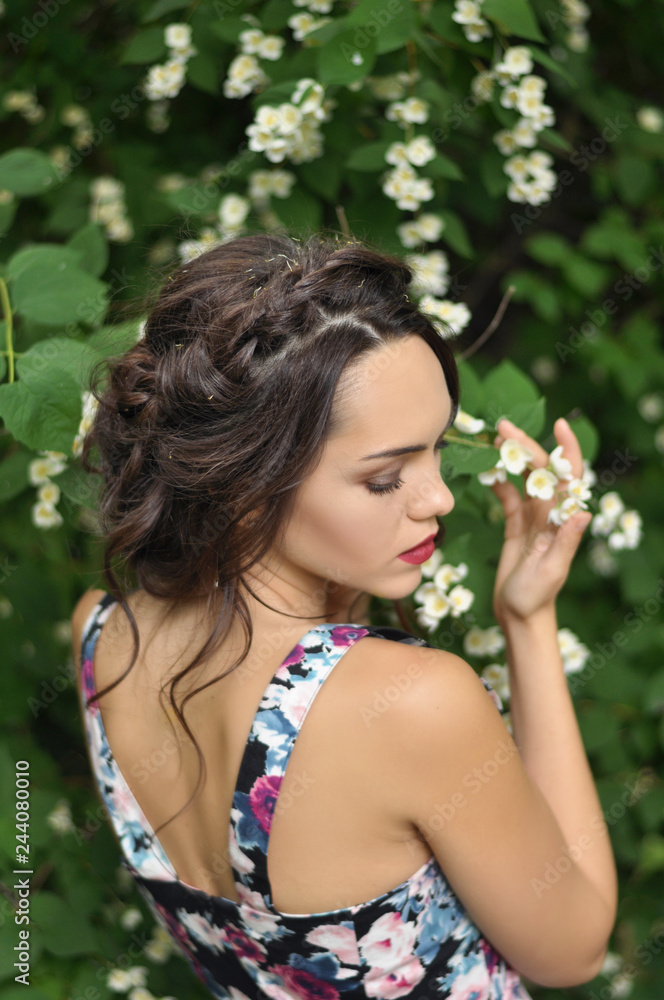 Young beautiful dark-haired woman in color dress with open shoulders in bright green blooming garden background Young beautiful dark-haired woman in color dress with open shoulders in bright green blooming garden background