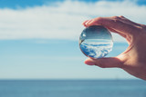 Woman's hand holding a crystal ball, looking through to the ocean and sky. Creative photography, crystal ball refraction