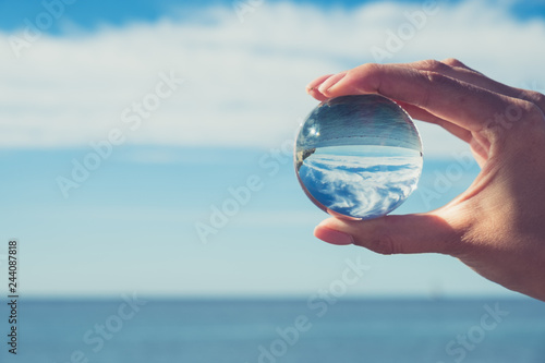 Woman's hand holding a crystal ball, looking through to the ocean and sky. Creative photography, crystal ball refraction