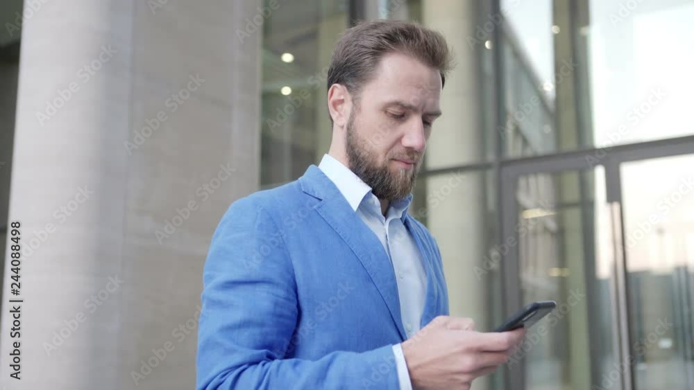 Panning shot of handsome bearded man standing outdoors and text messaging on his cell hone, looking away thoughtfully and smiling slightly Panning shot of handsome bearded man standing outdoors and text messaging on his cell hone, looking away thoughtfully and smiling slightly