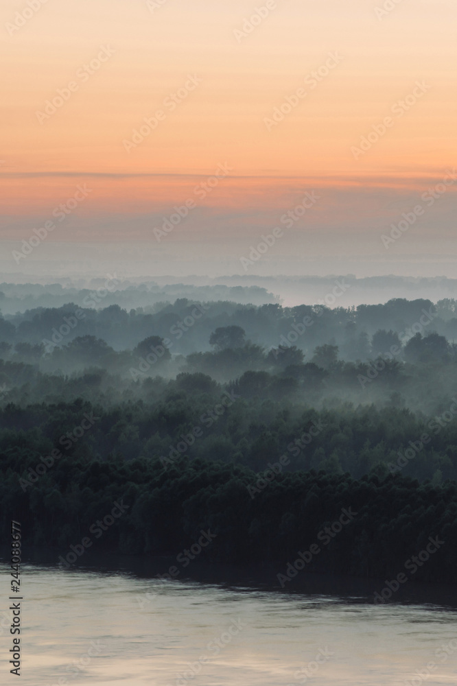 Mystical view on riverbank  of large island with forest under haze at early morning. Eerie mist among layers from tree silhouettes under predawn sky. Morning atmospheric landscape of majestic nature.