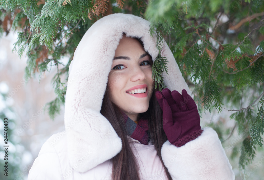 Obraz premium Portrait of young beautiful emotional woman in hooded down coat and gloves on snow covered garden background. Winter snowy landscape