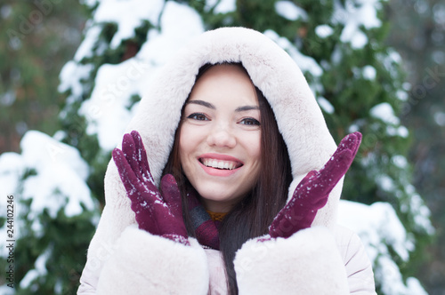 Portrait of young beautiful emotional woman in hooded down coat and gloves on snow covered garden background. Winter snowy landscape