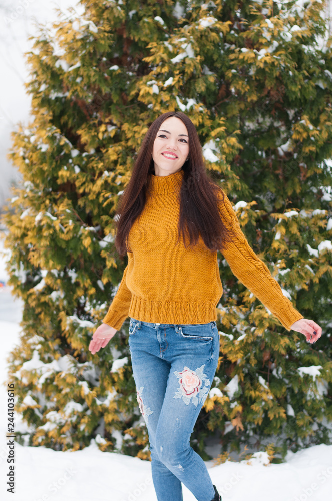 Portrait of young dark haired beautiful smiling woman in yellow knitted sweater and blue jeans on snow covered garden background. Winter snowy landscape Portrait of young dark haired beautiful smiling woman in yellow knitted sweater and blue jeans on snow covered garden background. Winter snowy landscape