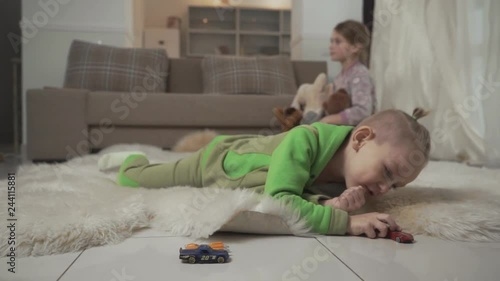 Little boy with stylish haircut plays with his toycars lying on the floor on fluffy carpet. Sister playing with a teddy bear on the background. photo